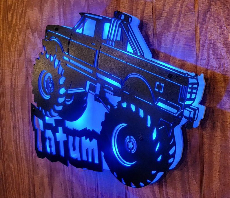 May include: A black metal silhouette of a monster truck with large tires and a blue light behind it. The name "Tatum" is written below the truck.