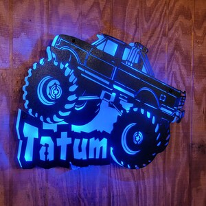 May include: A black metal silhouette of a monster truck with large tires and the name "Tatum" in blue lettering. The silhouette is lit from behind with blue light.