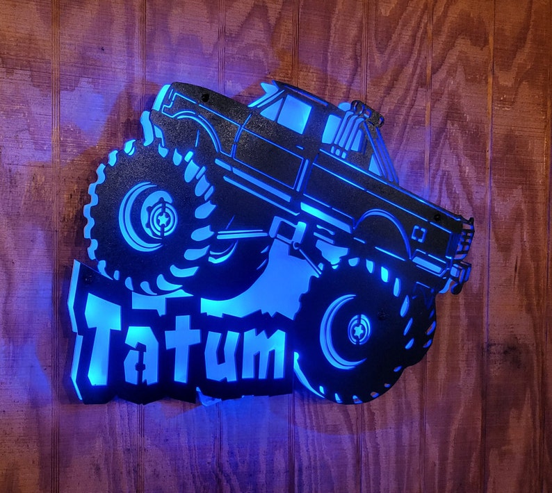 May include: A black metal silhouette of a monster truck with large tires and the name "Tatum" in blue lettering. The silhouette is lit from behind with blue light.