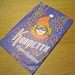 May include: A purple and white card with the text "Konfetti Karnaval'noe" in a stylized font. The card features a colorful illustration of a person wearing a jester hat and mask.