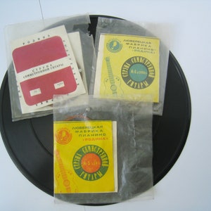May include: A collection of vintage guitar string packets and booklets, featuring Cyrillic text and a retro aesthetic. The packets are in clear plastic, with red and yellow labels. The items are on a black circular base.