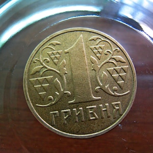 May include: A close-up of a bronze Ukrainian 1 Hryvnia coin. The coin features the number 1, decorative floral elements, and the word "ГРИВНЯ" in Cyrillic script. The coin is set against a blurred background.