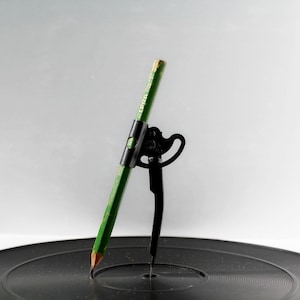 May include: A green pencil held by a black compass tool, positioned on a black vinyl record. The pencil is angled, with the point touching the record's surface. The compass has a decorative design.