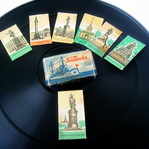 May include: A collection of vintage matchbox covers featuring illustrations of landmarks in Saint Petersburg, Russia. The matchboxes are arranged around a small blue box with the word "Baltika" printed on it. The matchboxes are on a black vinyl record.