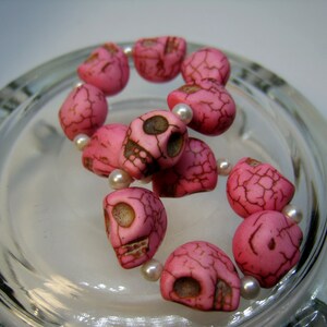 May include: Pink skull beads with white spacer beads strung on a bracelet. The beads are made of a textured material that resembles bone.