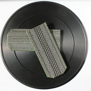 May include: Two rectangular, patterned objects with a green border rest on a black vinyl record. The objects have a geometric design in shades of gray and white. The record is black with concentric grooves.