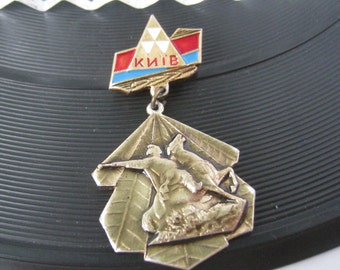 Puede incluir: Un pin de metal dorado con un emblema triangular rojo, blanco y azul que dice "КИЇВ" y una representación de una persona a caballo.