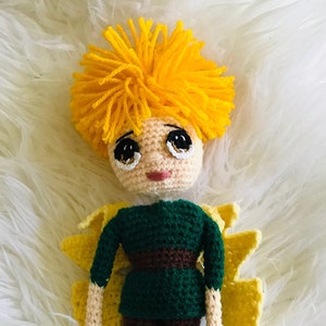 May include: A crocheted doll with yellow hair, a green shirt, brown pants, and yellow wings. The doll has a sweet expression with large eyes.