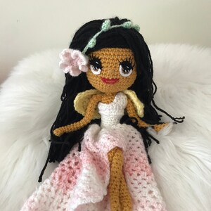 May include: Crocheted doll with brown skin, black hair, and a pink and white dress. The doll is wearing a flower crown and has large eyes.