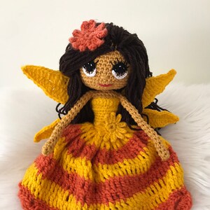 May include: A crocheted fairy doll with brown hair, a yellow and orange dress, and large eyes. The doll has wings and a flower on its head.
