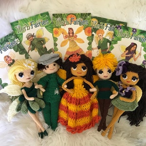 May include: Six crocheted dolls, each with a different color scheme and design. The dolls are dressed in green, orange, yellow, and brown. The dolls are surrounded by several books with the title "Herb Fairies" on the cover.