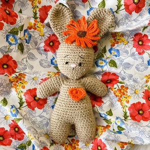 May include: A crocheted brown bunny with orange flower accents and a small heart on its chest. The bunny is lying on a floral fabric background.
