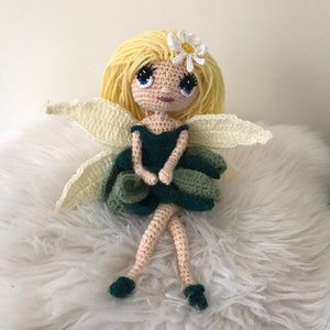 May include: A crocheted fairy doll with blonde hair, a green dress, and white wings. The doll is sitting on a white fluffy surface.