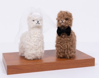 May include: Two alpaca figurines, one wearing a white veil and the other a black bow tie, stand on a wooden platform.