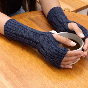 May include: A pair of gray fingerless gloves with a cable knit pattern. The gloves are worn by a person holding a white mug with a dark liquid inside.