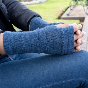 May include: A pair of blue knitted fingerless gloves. The gloves are a solid blue color with a ribbed design on the upper part of the glove. The person is wearing a black jacket and blue jeans.