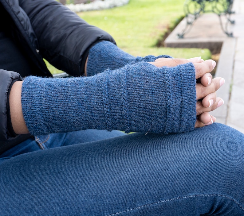 May include: A pair of blue knitted fingerless gloves. The gloves are a solid blue color with a ribbed design on the upper part of the glove. The person is wearing a black jacket and blue jeans.