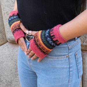 May include: A pair of pink fingerless gloves with a colorful geometric pattern. The gloves are made of knitted wool and have a brown leather strap.