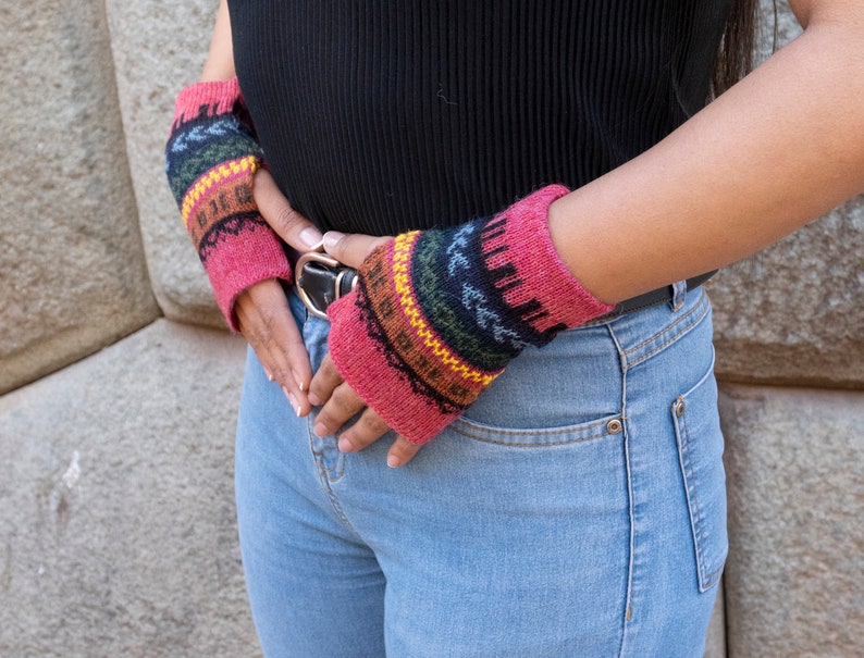 May include: A pair of pink fingerless gloves with a colorful geometric pattern. The gloves are made of knitted wool and have a brown leather strap.