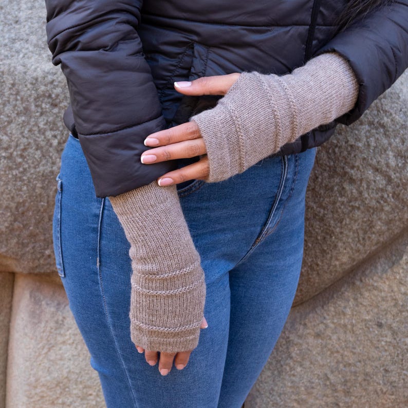 May include: Tan fingerless gloves with ribbed detailing. The gloves are made of a knitted material and are worn with a black jacket and blue jeans. The gloves are a neutral color and appear to be warm.