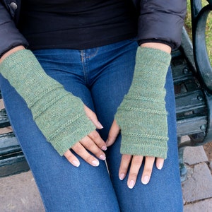 May include: A pair of green knitted fingerless gloves. The gloves are a muted green color with ribbed detailing around the wrist and upper arm. The gloves are worn by a person sitting on a park bench.