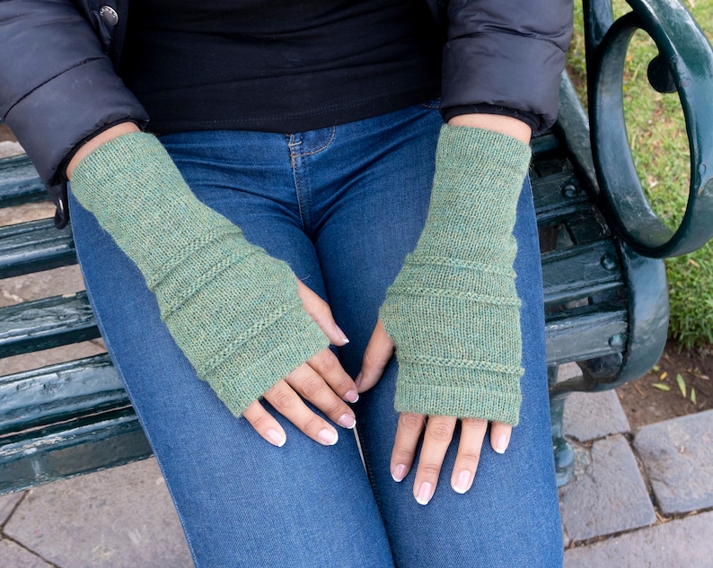 May include: A pair of green knitted fingerless gloves. The gloves are a muted green color with ribbed detailing around the wrist and upper arm. The gloves are worn by a person sitting on a park bench.