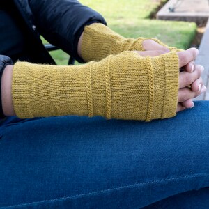 May include: Mustard yellow fingerless gloves with ribbed detailing. The gloves are made of a knitted material and cover the hands up to the knuckles. The person is wearing blue jeans and a black jacket. The gloves are a warm accessory.