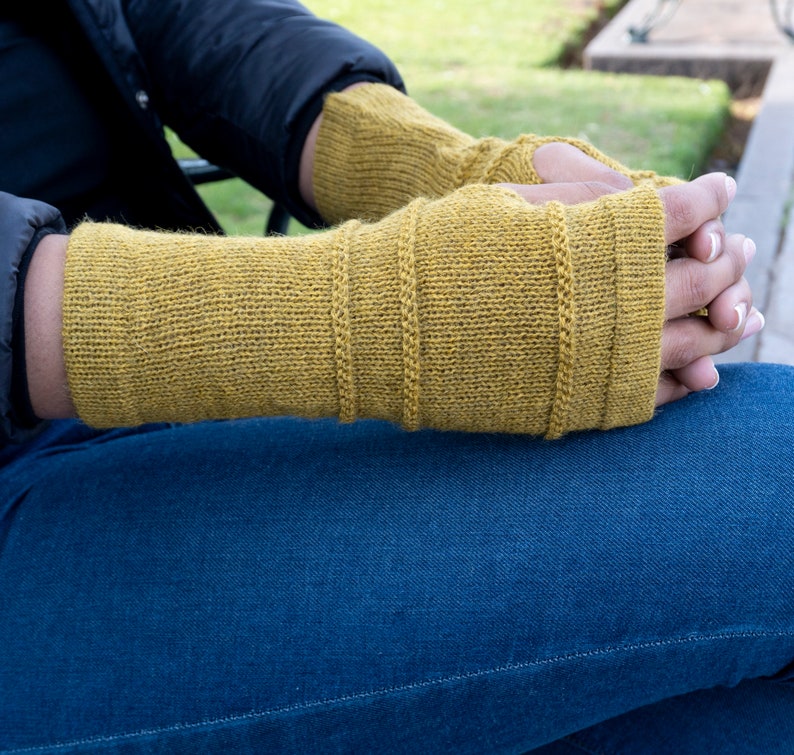 May include: Mustard yellow fingerless gloves with ribbed detailing. The gloves are made of a knitted material and cover the hands up to the knuckles. The person is wearing blue jeans and a black jacket. The gloves are a warm accessory.