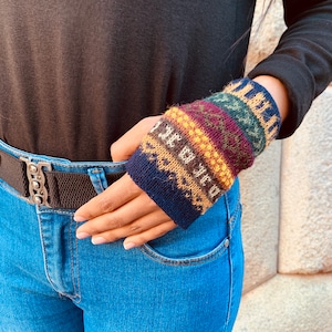 May include: A close-up of a person's hand wearing a colorful knitted wrist warmer with a blue, brown, yellow, red, and green pattern.