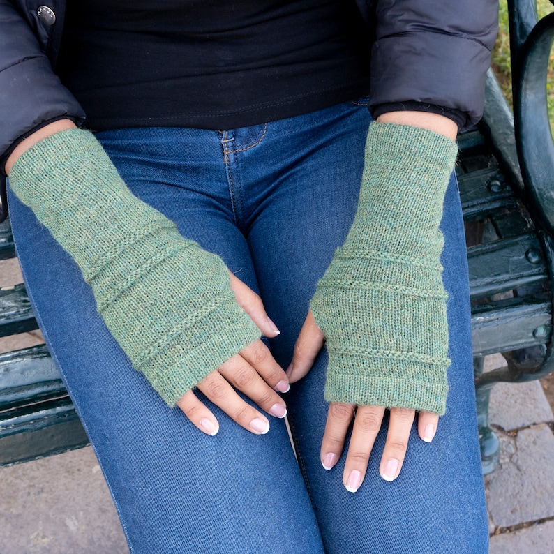 May include: A pair of green knitted fingerless gloves. The gloves are a muted green color with horizontal ribbed detailing. The gloves are designed to cover the hand and wrist, leaving the fingers exposed. The person is wearing blue jeans and a black jacket.