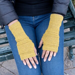 May include: Mustard yellow knitted fingerless gloves. The gloves have a ribbed design at the wrist and mid-arm, and are made of a soft, textured yarn. The gloves are shown on a person wearing a black jacket and blue jeans.