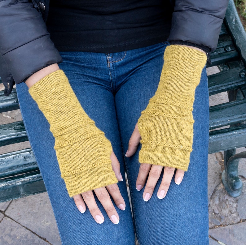 May include: Mustard yellow knitted fingerless gloves. The gloves have a ribbed design at the wrist and mid-arm, and are made of a soft, textured yarn. The gloves are shown on a person wearing a black jacket and blue jeans.