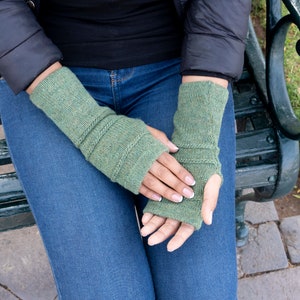 May include: Green knitted fingerless gloves with ribbed detailing. The gloves are a muted green color and appear to be made of a soft, textured yarn. The design includes ribbed cuffs and a section of horizontal ribbing near the wrist.