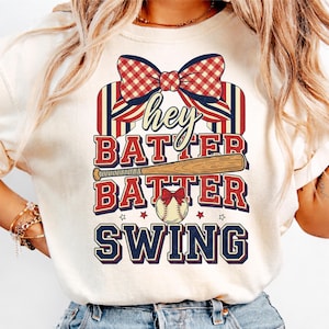 Può includere: T-shirt color crema con grafica a tema baseball. Il design presenta le parole "Hey Batter Batter Swing" in rosso e blu navy, insieme a una mazza da baseball, una palla e un fiocco.