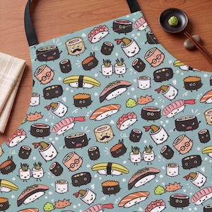 May include: A light blue apron featuring a repeating pattern of cartoon sushi pieces with faces. The design includes various types of sushi rolls, nigiri, and shrimp tempura, some with glasses or mustaches, scattered across the fabric with small white hearts.