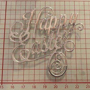 May include: Silver-coloured metal cutout of the words "Happy Easter" with a decorative swirl at the end of the "r" in Easter.