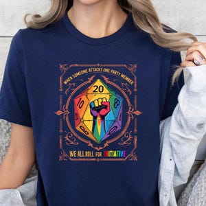 May include: Navy blue t-shirt featuring a colorful D20 die graphic with a fist and the text "WHEN SOMEONE ATTACKS ONE PARTY MEMBER WE ALL ROLL FOR INITIATIVE". The graphic is in a decorative frame.
