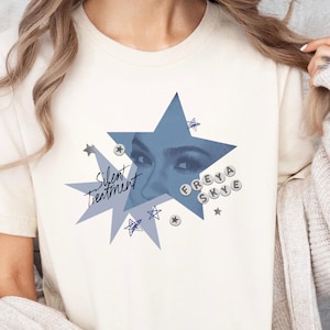 May include: Cream-colored t-shirt featuring a blue starburst design with the text "Silent Treatment" and "Freya Skye." The design includes a close-up of eyes within the star. Small star graphics surround the main design. A casual, everyday tee.
