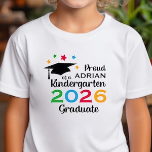 May include: White t-shirt with a colorful graphic. The graphic includes a black graduation cap, stars, and the text "Proud of a ADRIAN Kindergarten 2026 Graduate" in various colors. The shirt is short-sleeved.
