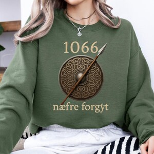 Może przedstawiać: Zielona bluza z numerem "1066" i napisem "n&aelig;fre forg&yacute;t". Projekt zawiera tarczę i wł&oacute;cznię. Tarcza ma brązowy celtycki wz&oacute;r węzła.