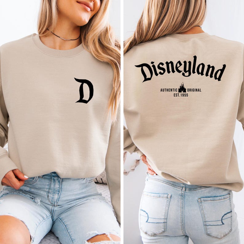 May include: Beige sweatshirt with a black "D" on the front. The back features the word "Disneyland" in a stylized font, with "AUTHENTIC ORIGINAL EST. 1955" below it. The sweatshirt is a neutral color, suitable for various outfits.