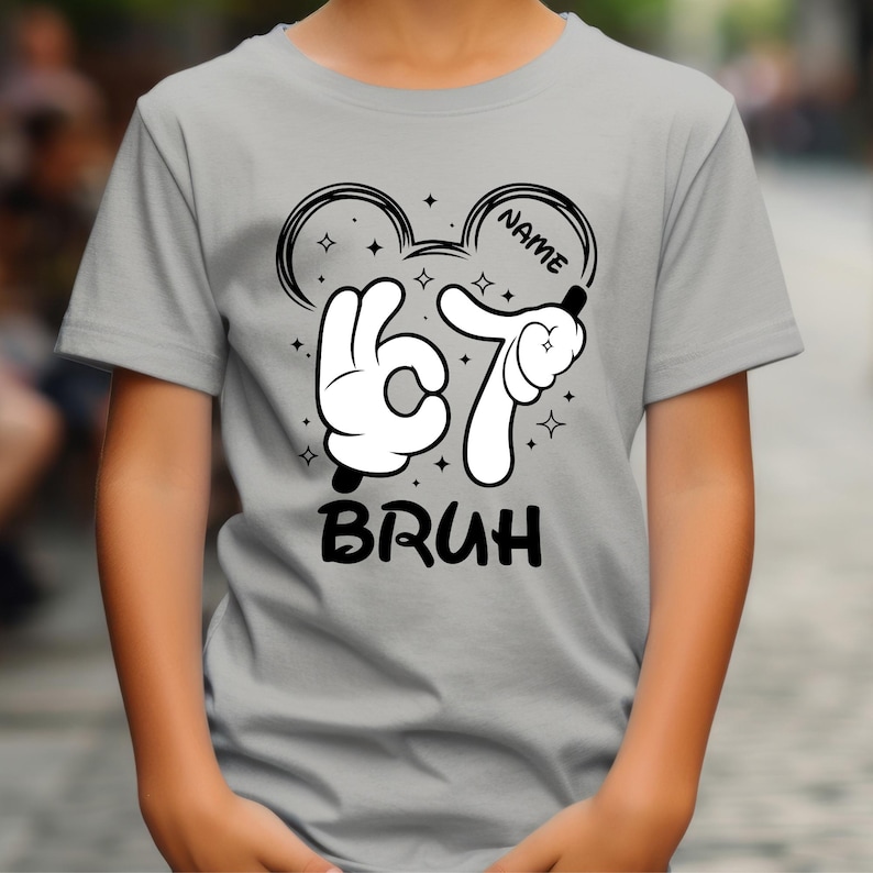 Puede incluir: Una camiseta gris claro con un dise&ntilde;o gr&aacute;fico en blanco y negro. El dise&ntilde;o incluye manos al estilo de Mickey Mouse formando el n&uacute;mero 67, la palabra "BRUH" y la palabra "NAME" dentro de una forma de oreja de Mickey Mouse. Peque&ntilde;as estrellas est&aacute;n dispersas alrededor del dise&ntilde;o.