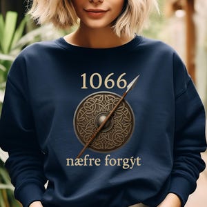Może przedstawiać: Granatowa bluza z nadrukiem roku "1066" nad wzorem tarczy i wł&oacute;czni. Tarcza ma wz&oacute;r celtyckiego węzła. Tekst "n&aelig;fre forg&yacute;t" znajduje się poniżej. Bluza jest w jednolitym kolorze.