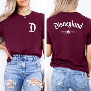 May include: Burgundy t-shirt with white Disneyland logo on the back and a white "D" on the front. The back of the shirt also has the text "AUTHENTIC ORIGINAL EST. 1955". The shirt is paired with blue jeans.