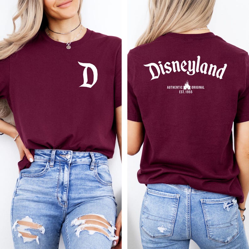 May include: Burgundy t-shirt with white Disneyland logo on the back and a white "D" on the front. The back of the shirt also has the text "AUTHENTIC ORIGINAL EST. 1955". The shirt is paired with blue jeans.