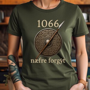 Może przedstawiać: Zielona koszulka z oliwkowym nadrukiem z napisem "1066" nad tarczą i wł&oacute;cznią. Tarcza ma wz&oacute;r celtyckiego węzła. Pod spodem znajduje się napis "n&aelig;fre forg&yacute;t". Koszulka wykonana jest z miękkiego materiału.