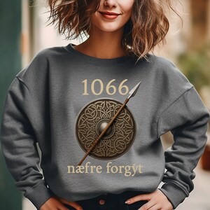 Może przedstawiać: Szara bluza z numerem "1066" nad wzorem tarczy i wł&oacute;czni. Tarcza jest okrągła ze skomplikowanym wzorem, a wł&oacute;cznia jest ustawiona po przekątnej. Tekst "n&aelig;fre forgyt" znajduje się poniżej.