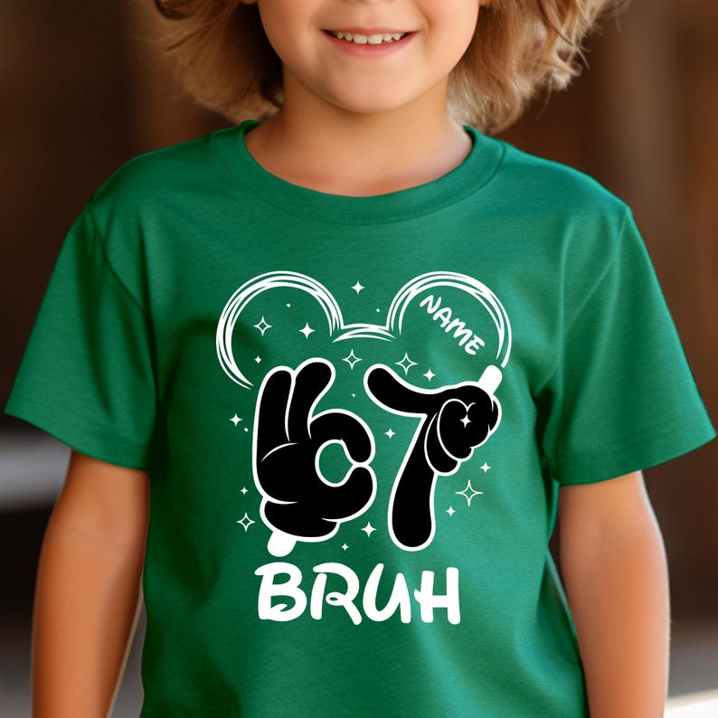 Puede incluir: Una camiseta verde con un dise&ntilde;o gr&aacute;fico blanco. El dise&ntilde;o incluye el n&uacute;mero "67" en un estilo de mano de dibujos animados, la palabra "BRUH" y un espacio para un nombre. El dise&ntilde;o est&aacute; rodeado de estrellas y orejas de Mickey Mouse.