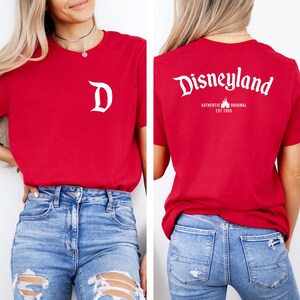 May include: Red t-shirt with a white "D" on the front and "Disneyland" printed on the back. The back also features "AUTHENTIC ORIGINAL EST. 1955". The shirt is a classic crew neck style.