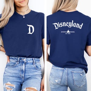 May include: Navy blue t-shirt with a white "D" on the front and "Disneyland" with "AUTHENTIC ORIGINAL EST. 1955" on the back. The shirt is paired with light blue denim jeans.
